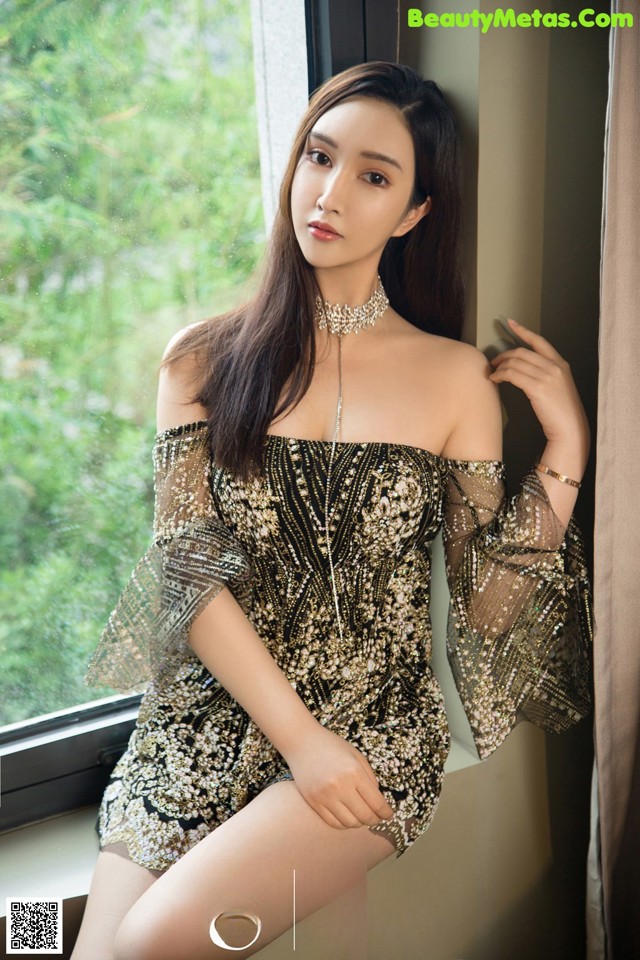 A woman in a black and gold dress sitting by a window.